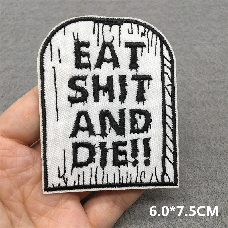 Embroidered patch with text 'EAT SHIT AND DIE!!' held by a hand on a gray background.
