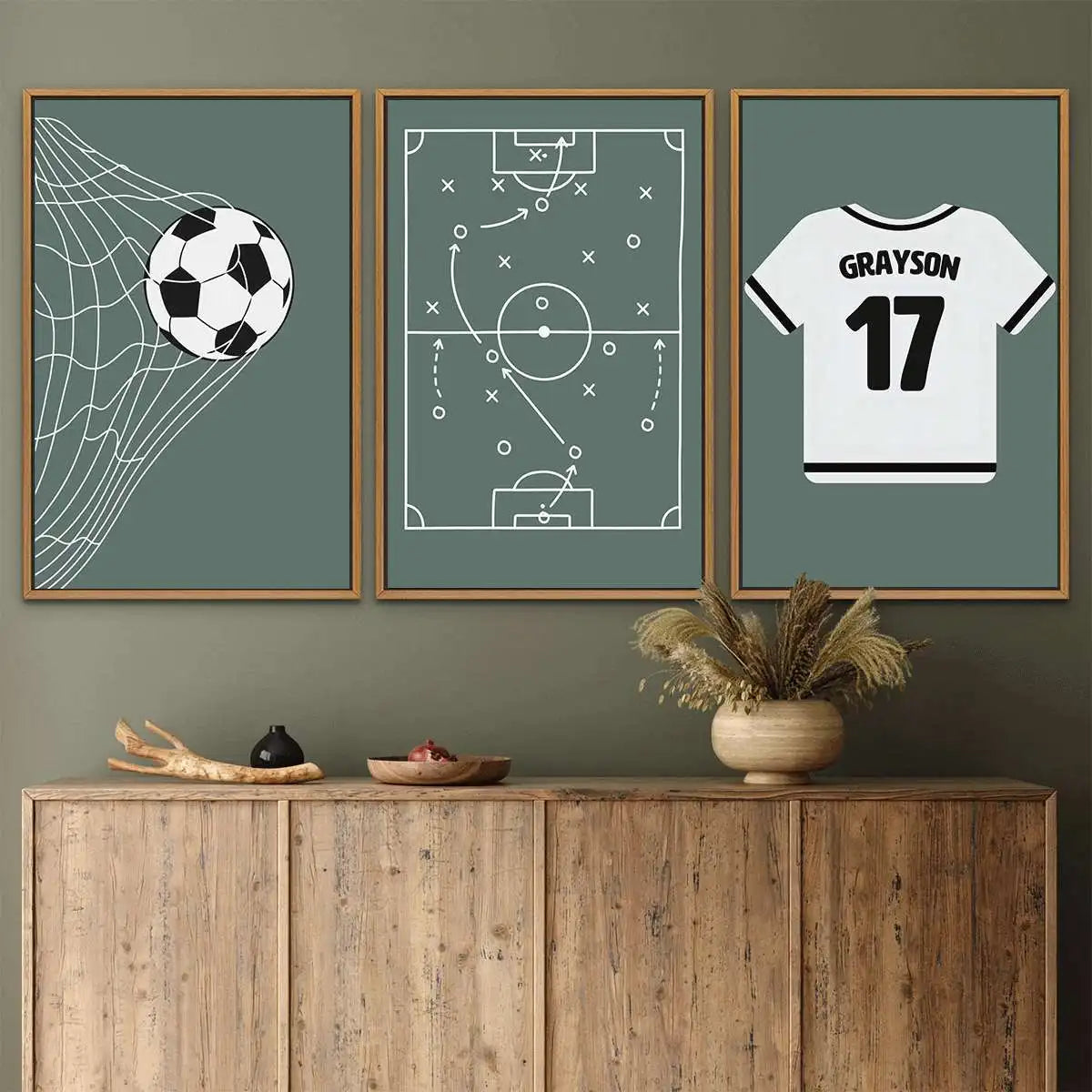 Three framed wall art prints with sports-themed designs hang above a wooden cabinet in a styled room. The left print shows a soccer ball net, the center shows a football field tactic diagram, and the right print shows a white sports jersey with the name GRAYSON and number 17.