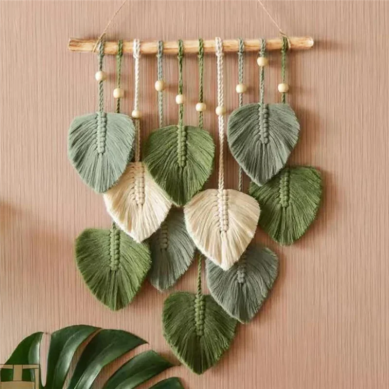Macrame wall hanging with leaf-shaped green and beige elements on a wooden stick against a beige wall.