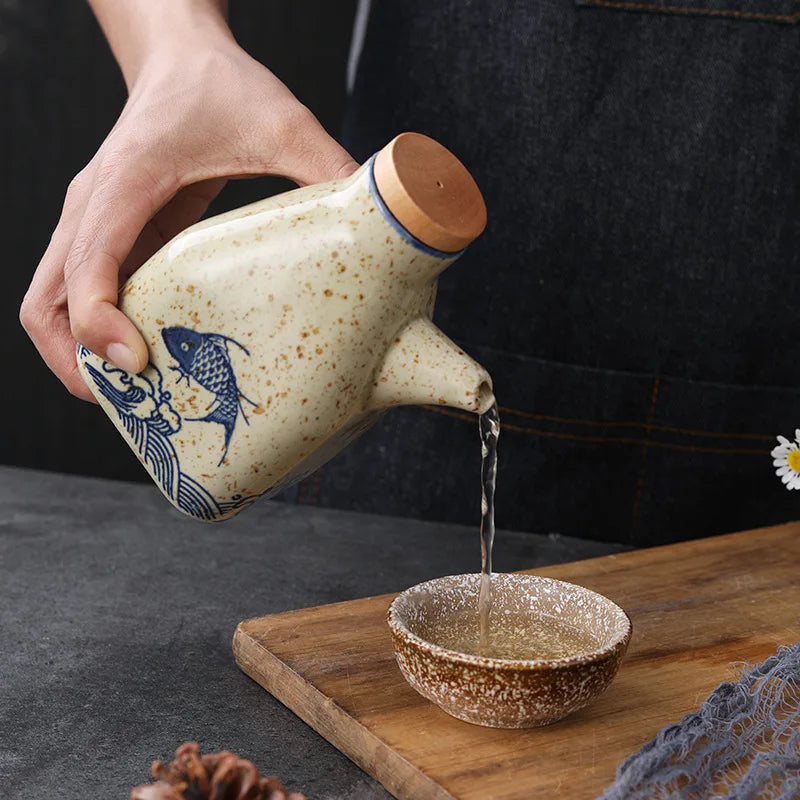 Person pouring liquid from a ceramic teapot with fish design into a small bowl on a wooden tray.