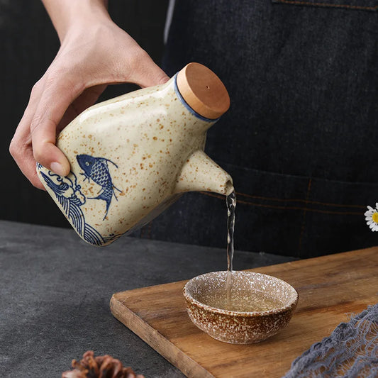 Person pouring liquid from a ceramic teapot with fish design into a small bowl on a wooden tray.