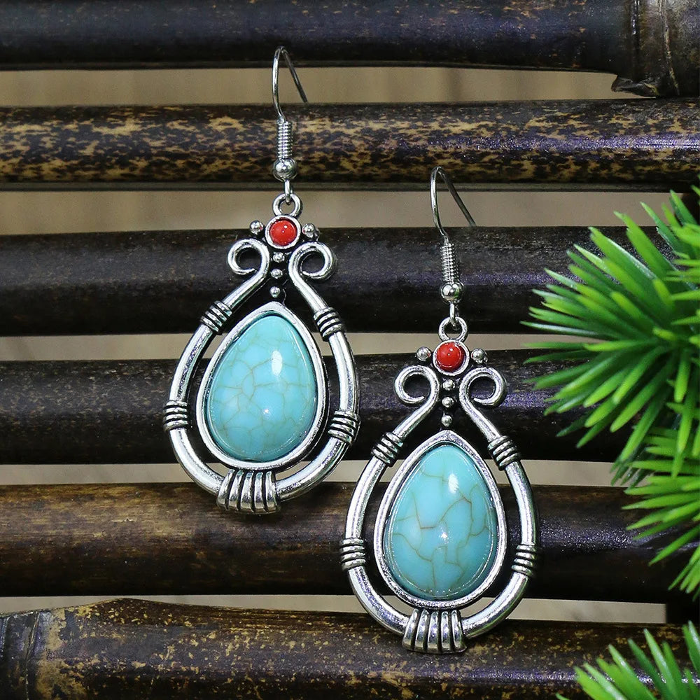 Turquoise teardrop earrings with silver and red accents on a wooden surface with green leaves.
