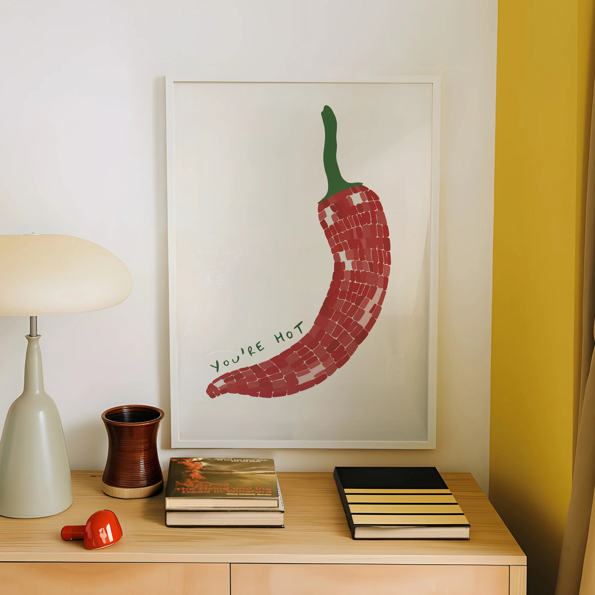 Framed artwork of a red chili pepper with 'You're Hot' text on a wall above a wooden shelf with books and decor items.