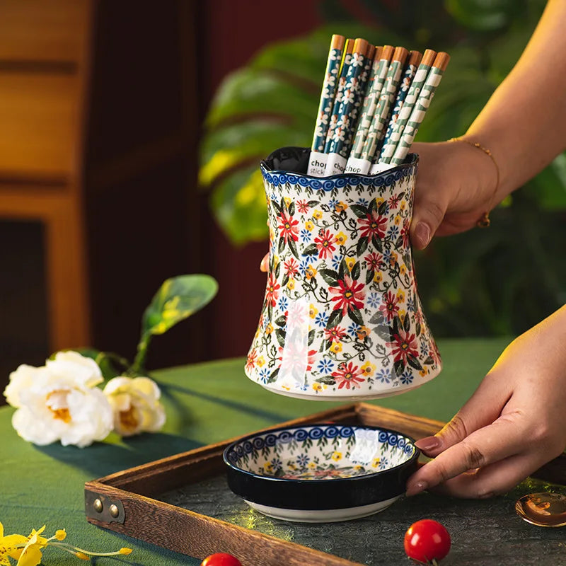 Decorative floral utensil holder with pens on a table with flowers and a plant in the background