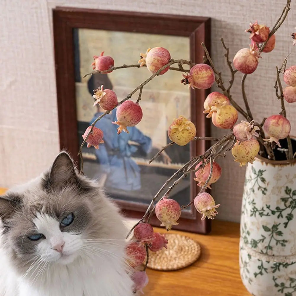 Cat sitting next to a decorative branch with fruit-like ornaments on a wooden surface.