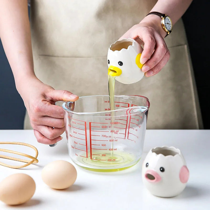 Person using a cute duck-shaped egg white separator to pour liquid into a measuring cup with a whisk and eggs on a table.
