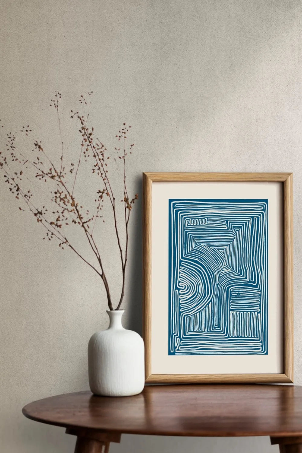 Framed abstract artwork on a wooden table with a white vase containing dried branches.