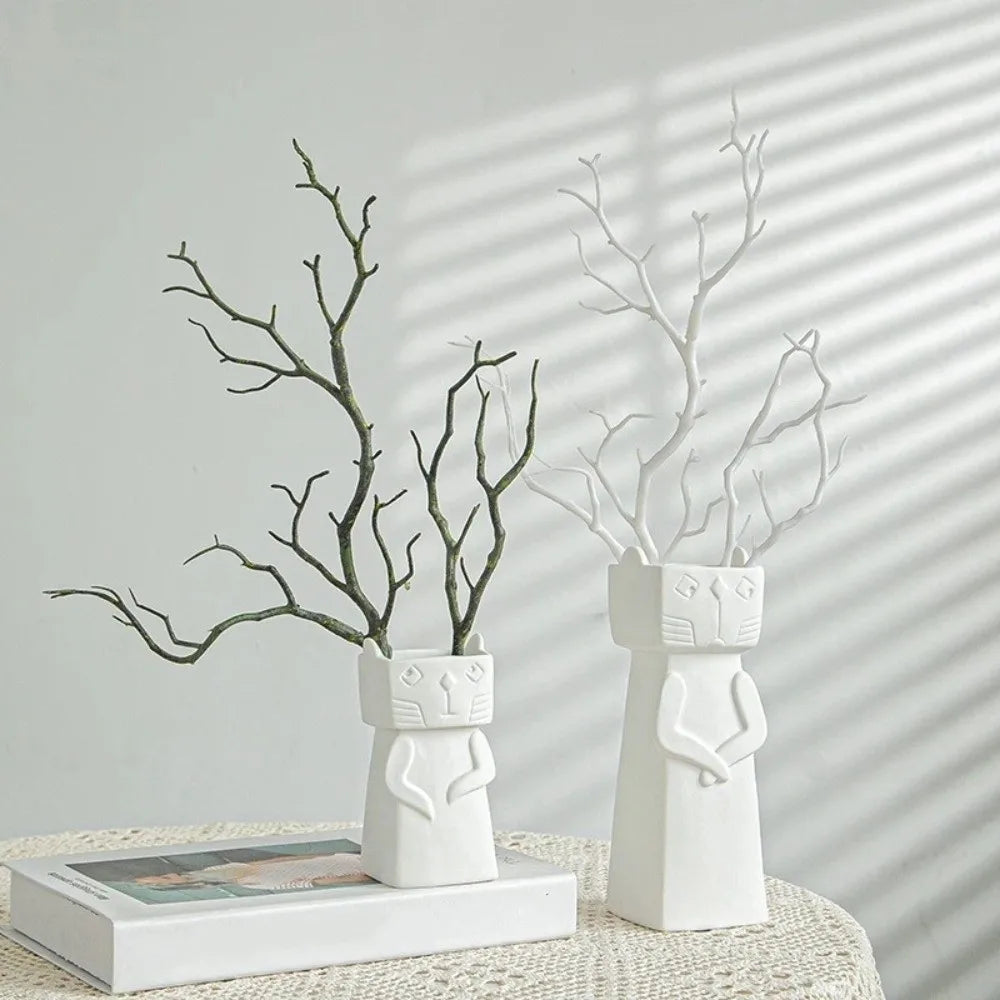 Two white ceramic cat-shaped vases with branches inside, displayed on a white textured tablecloth with soft window shadow patterns on a light gray wall.
