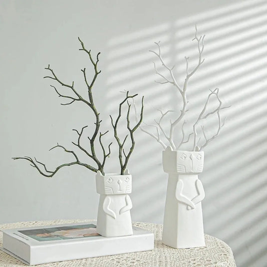 Two white ceramic cat-shaped vases with branches inside, displayed on a white textured tablecloth with soft window shadow patterns on a light gray wall.