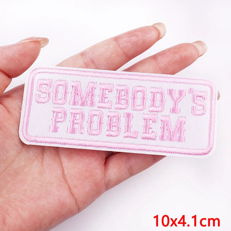 Hand holding a pink patch with 'SOMEBODY'S PROBLEM' text on a white background
