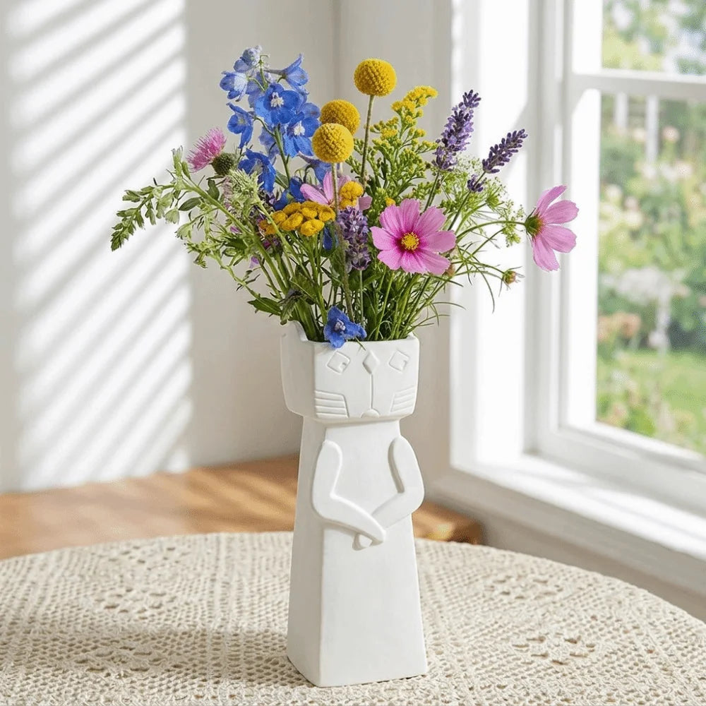White minimalist cat ceramic vase filled with colourful wildflowers and displayed on a table beside a bright window.