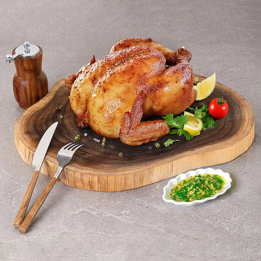 Roasted chicken on a wooden cutting board with garnishes and a small dish of green sauce.