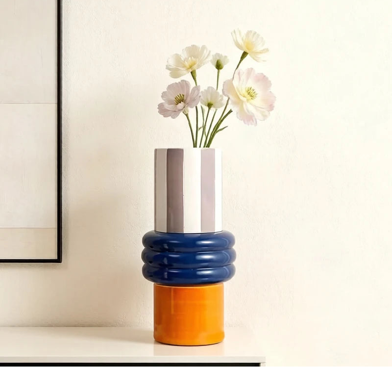 A modern stacked ceramic vase with a white and gray striped top, a ribbed navy blue middle section, and a bright orange base, holding white flowers on a white shelf against a beige wall.
