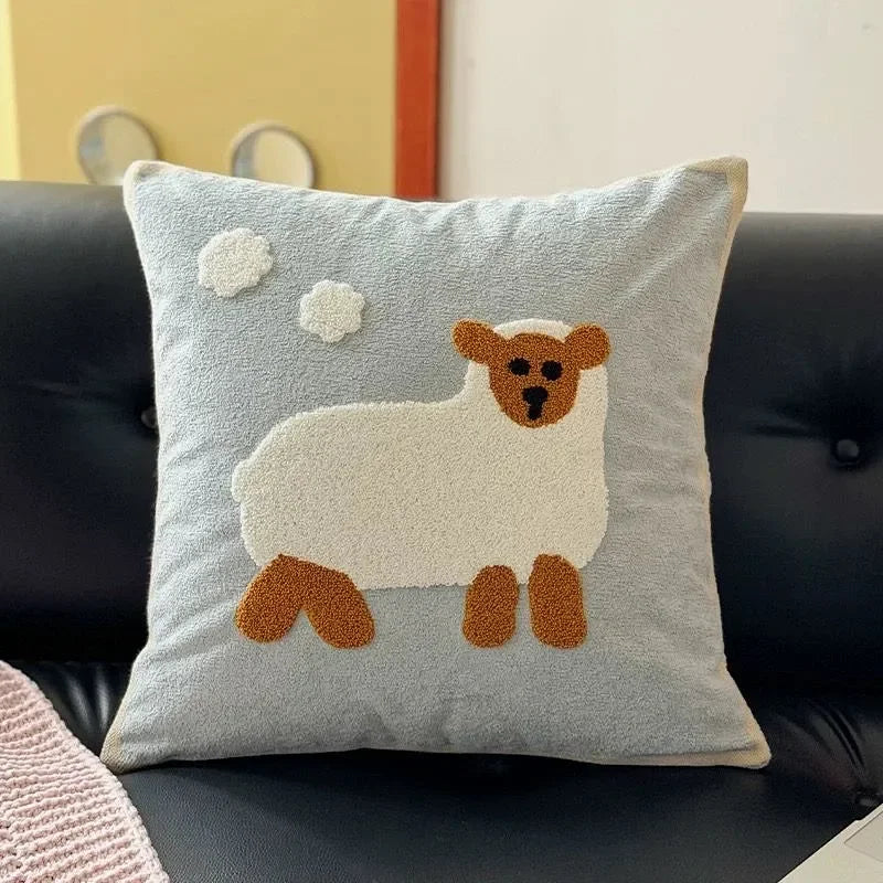 A square throw pillow with a light blue background featuring a raised, textured white sheep and two white clouds, displayed on a black leather sofa.