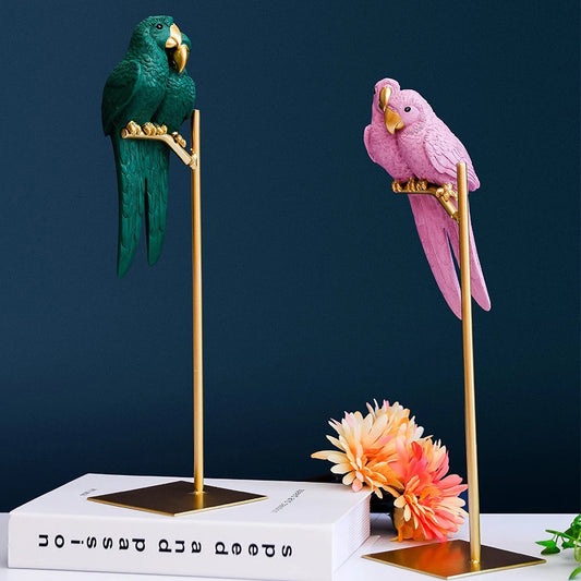 Two parrot figurines, one green and one pink, on gold stands with a dark blue background.