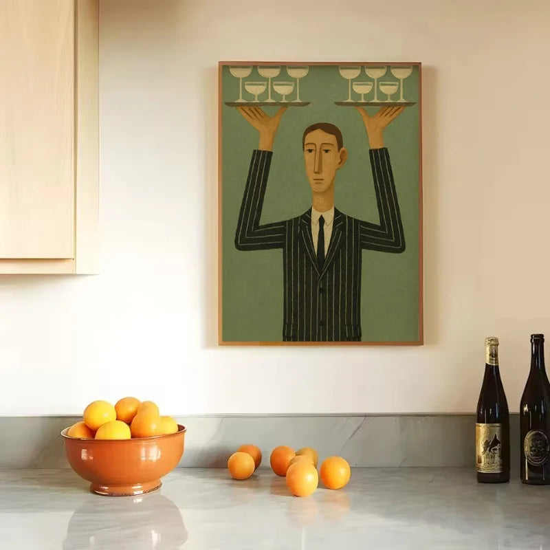 Framed artwork of a man balancing wine glasses on a kitchen counter with oranges and wine bottles.
