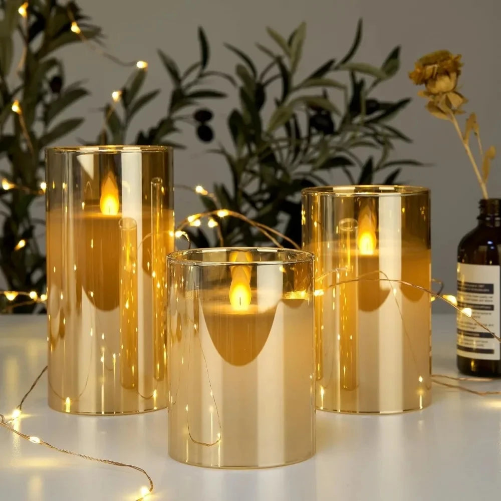 Three gold candle holders with lit candles on a surface with plants and a bottle in the background.