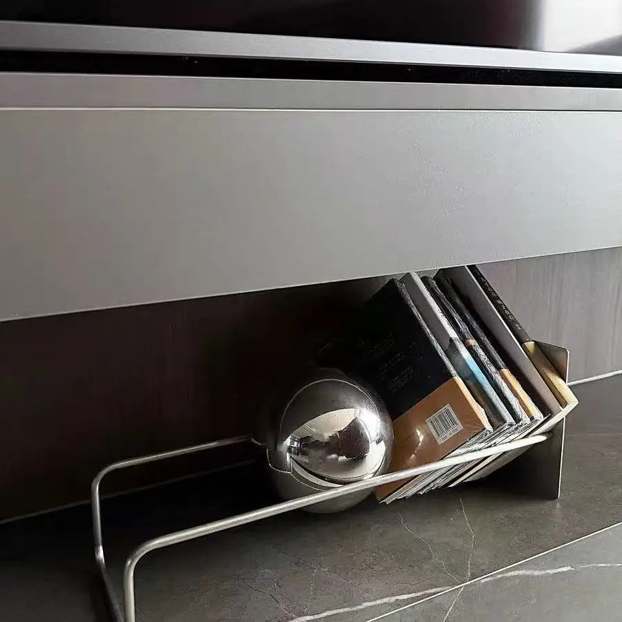 A modern silver metal bookend placed underneath a white entertainment console on a dark gray tiled floor, holding a stack of DVDs and a reflective silver sphere.