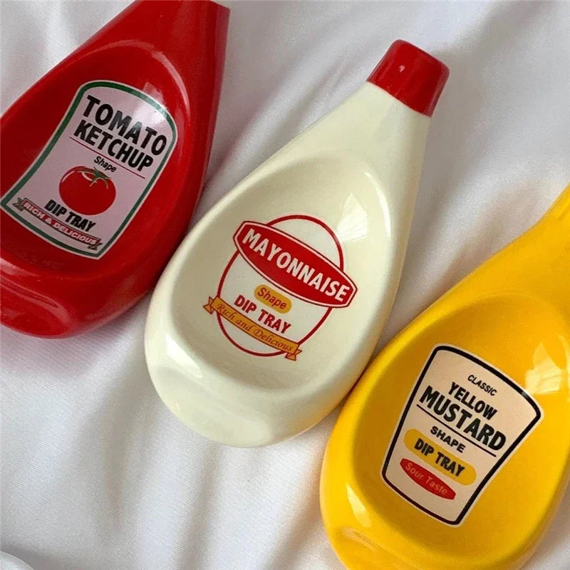 Three ceramic dip trays shaped like ketchup, mayonnaise, and mustard, arranged on a white fabric background.