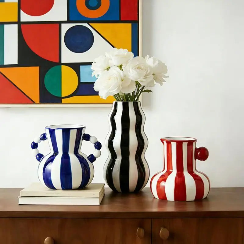 Three decorative ceramic vases with bold vertical stripes and sculptural forms displayed on a wooden cabinet. The left vase is blue and white with two handles and sits on books; the center vase is black and white with wavy vertical stripes and holds white flowers; the right vase is red and white with a side handle.