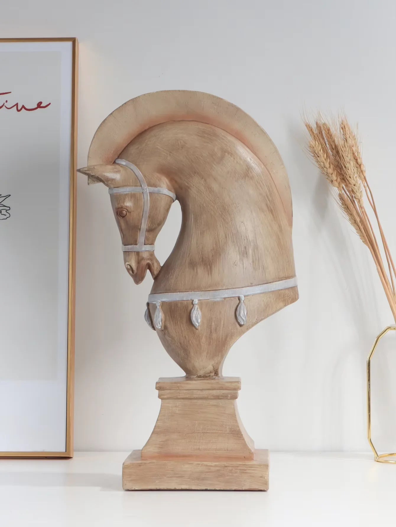 Wooden horse head sculpture on a white surface with a framed picture and dried plants in the background.
