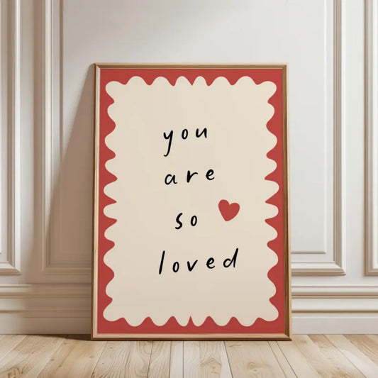 Framed artwork with 'you are so loved' text and a heart on a beige wall.