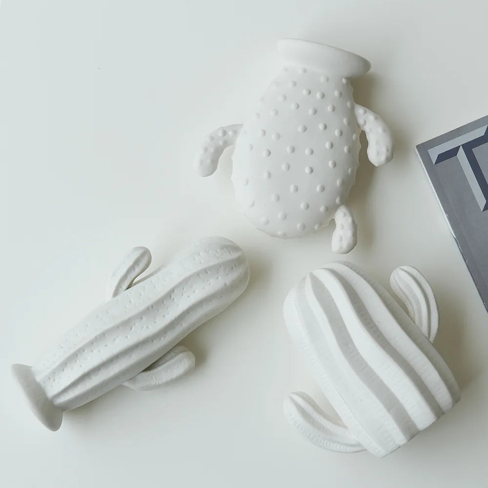 Three white ceramic cactus-shaped decorative objects with different textured and striped patterns, arranged on a clean white surface next to a gray book corner.
