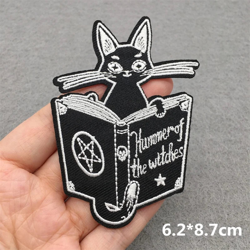 Embroidered patch of a black cat reading a book with a pentagram, held by a hand on a gray background.