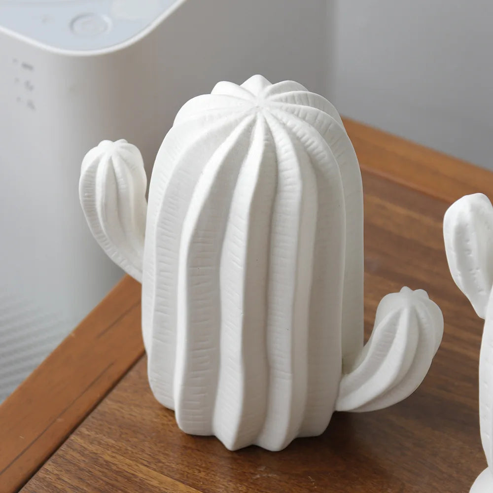 A white cactus-shaped decorative object with ribbed vertical texture and small side arms, shown on a wooden tabletop with a blurred indoor background.