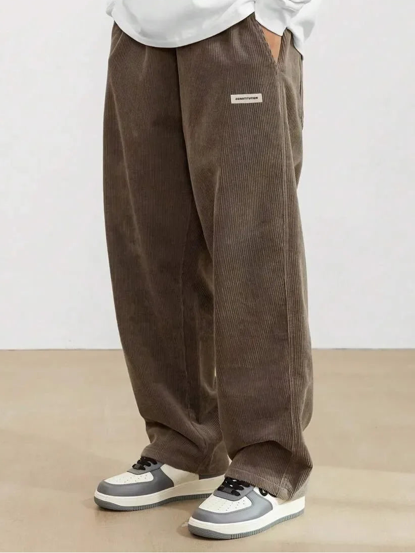 Person wearing brown corduroy pants with a white shirt and gray sneakers on a plain background, featuring the brand 'YIGUDAR'.