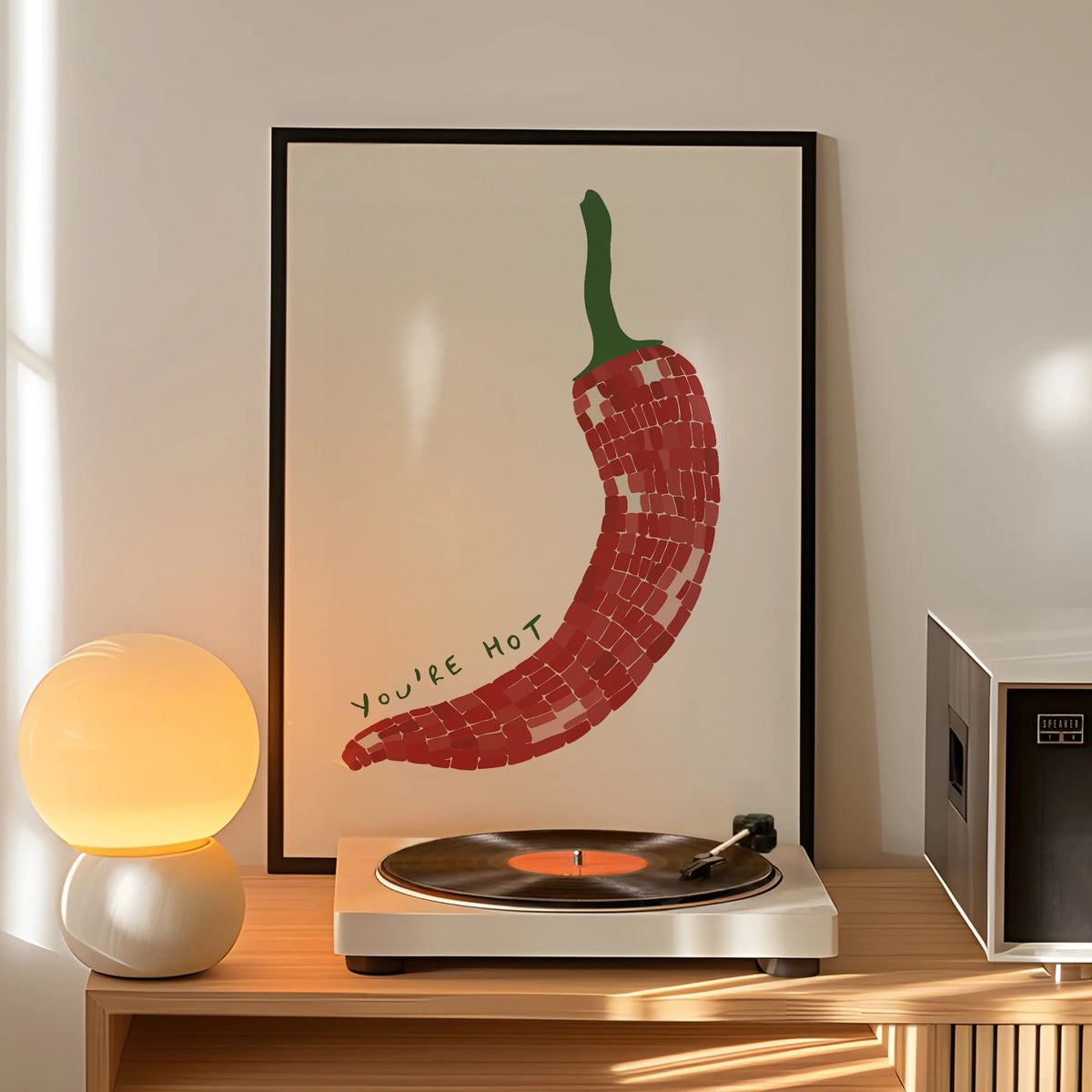 Framed artwork of a red chili pepper with 'You're Hot' text above a record player on a wooden shelf.