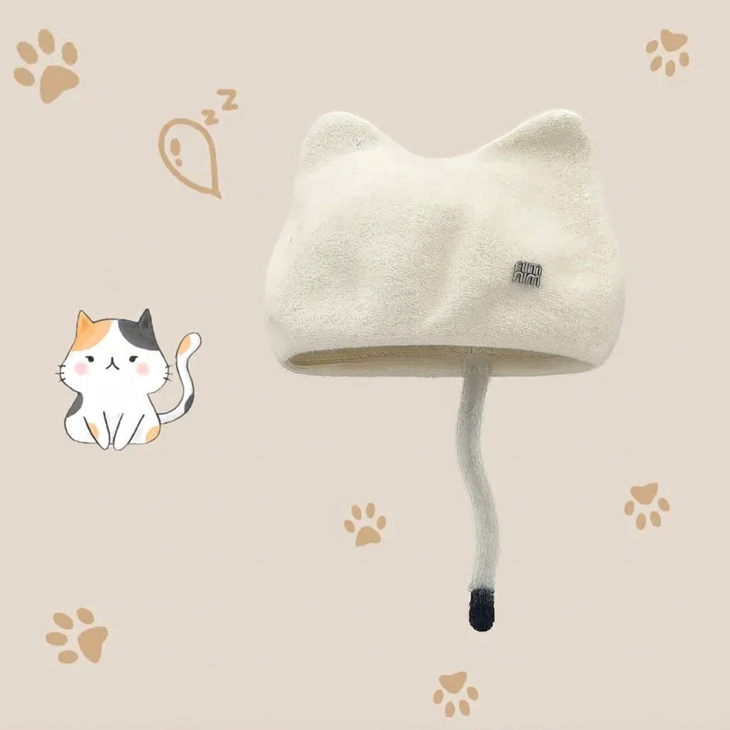 A cream-colored cat-ear wool hat with a long tail detail and a small square logo patch, shown on a beige background with paw prints and a cartoon cat illustration. 
