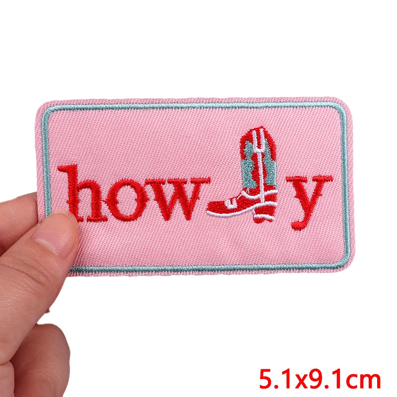 Pink patch with 'howdy' text and boot design held by a hand, on a white background.