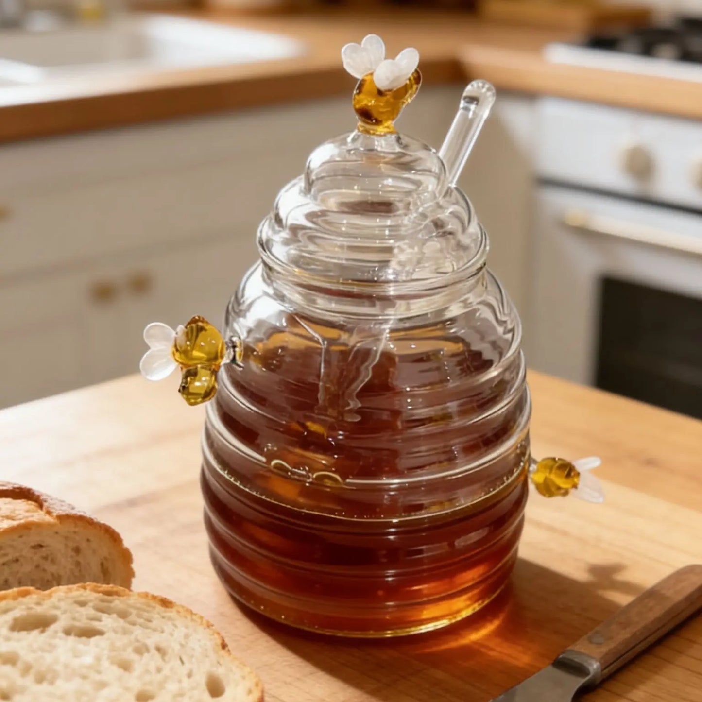 Glass honey container with a bee design on a kitchen counter with bread and a knife.