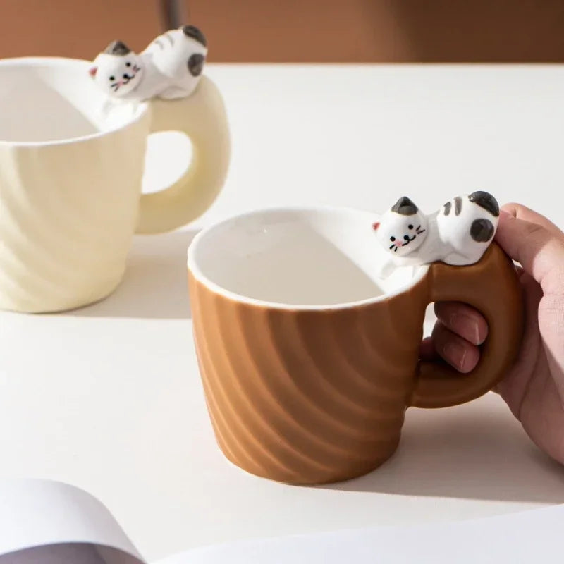 Brown mug with a cat-shaped handle held by a hand, with another similar mug in the background.