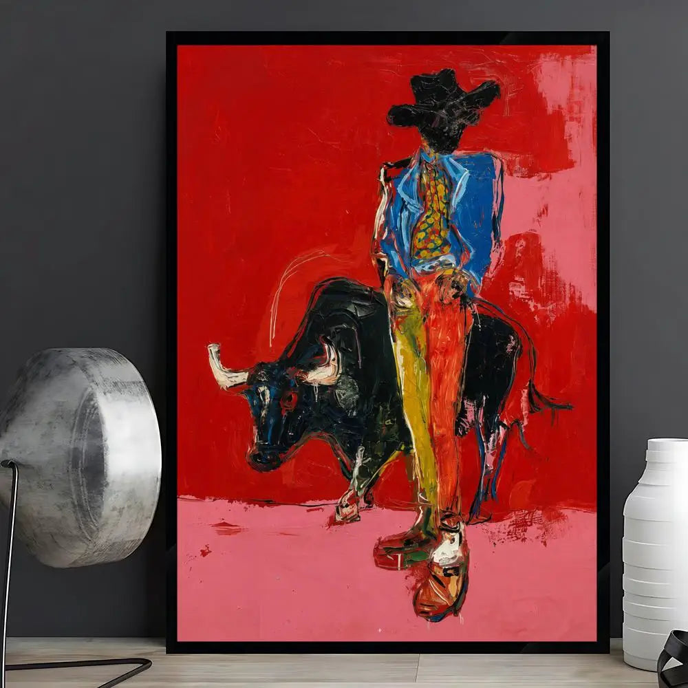 A framed abstract art print depicting a bullfighter in a blue jacket and yellow trousers facing a black bull, set against a vibrant red and pink background. The framed print is displayed on a light wood floor against a dark gray wall, flanked by a silver lamp on the left and a white ceramic vase on the right.