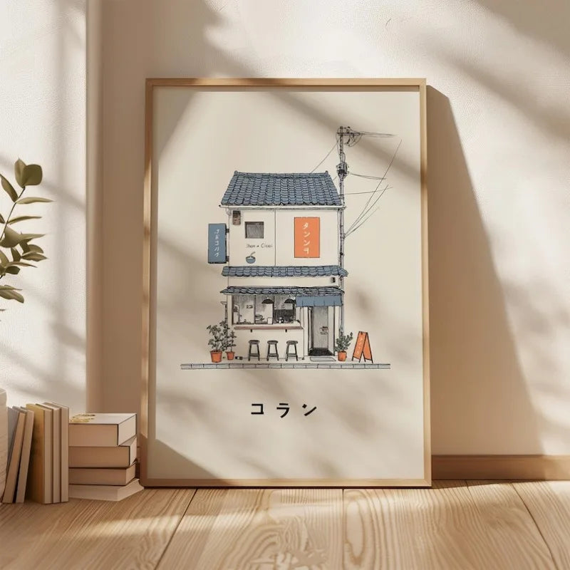 A framed art print featuring a hand-drawn illustration of a small Japanese-style house and cafe, displayed on a wooden floor against a beige wall with soft window shadows.