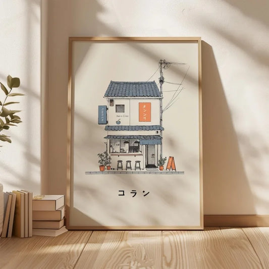 A framed art print featuring a hand-drawn illustration of a small Japanese-style house and cafe, displayed on a wooden floor against a beige wall with soft window shadows.