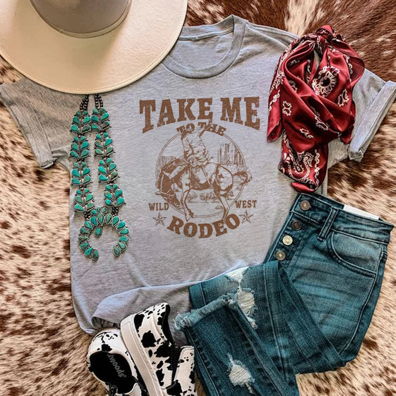 Gray t-shirt with 'Take Me to the Wild West Rodeo' design, cowboy hat, necklace, bandana, and jeans on a fur surface.