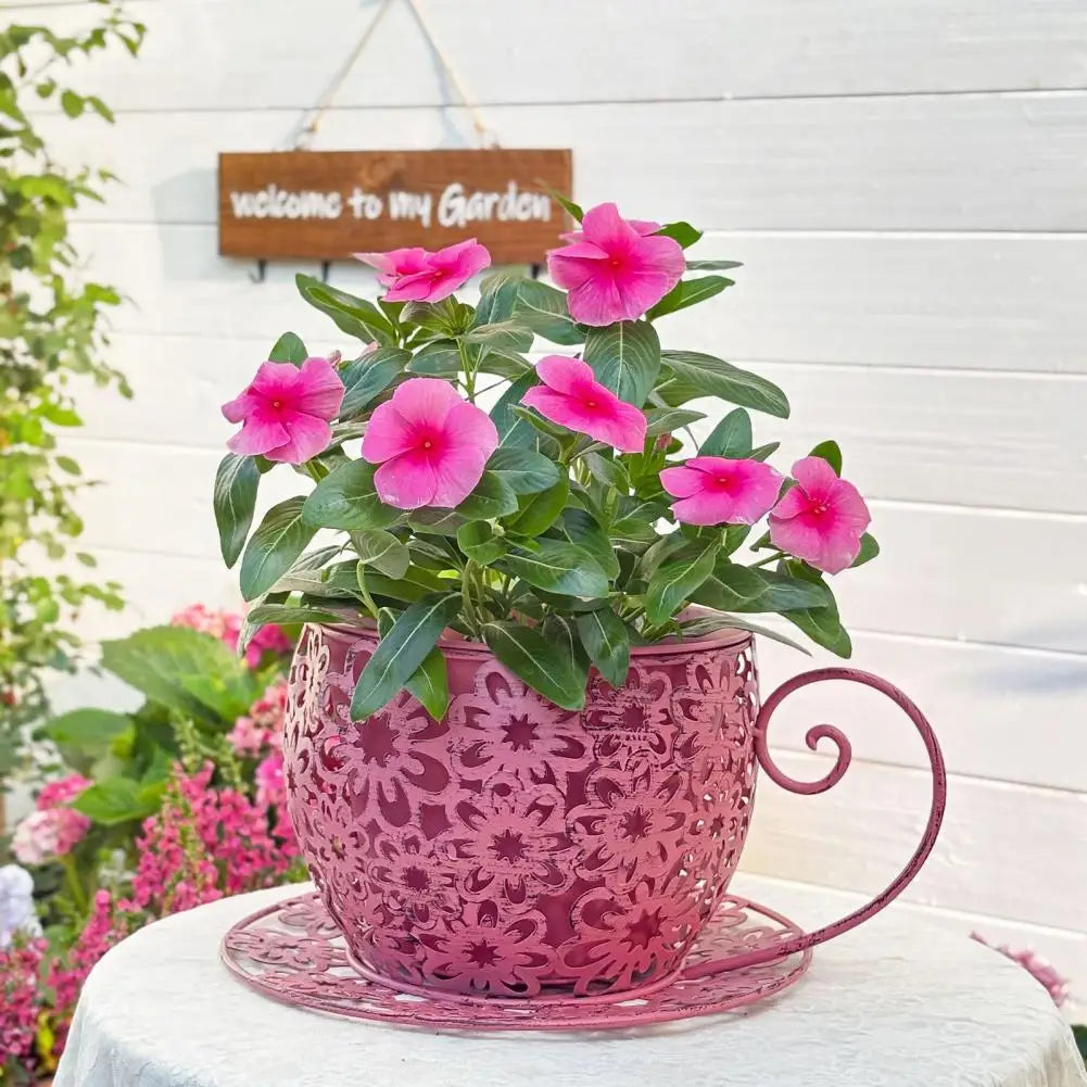 Pink flowers in a decorative pink pot on a white surface with a 'welcome to my garden' sign in the background.
