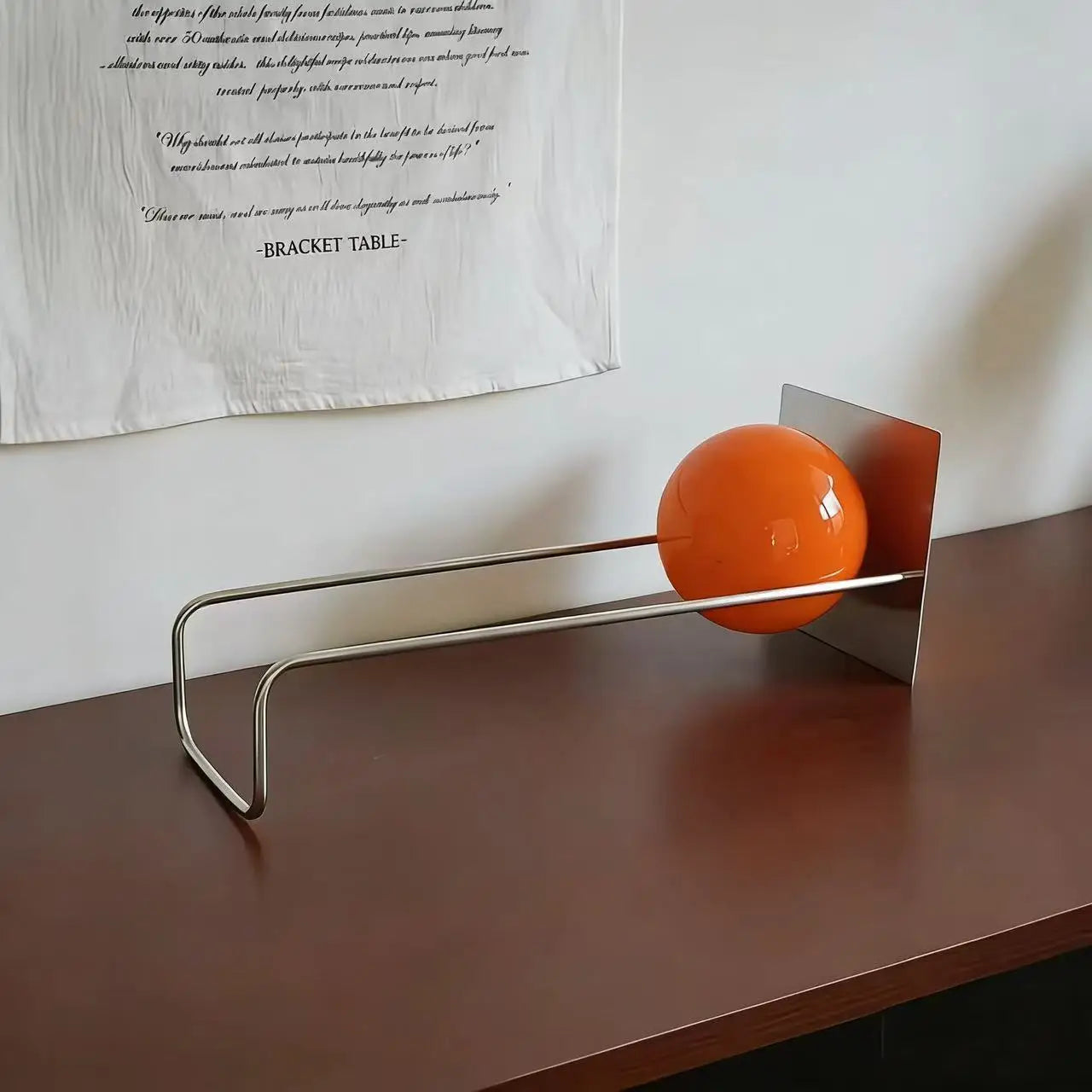 A modern orange glass sphere A modern metal bookend , set on a dark wooden table against a white wall with a hanging fabric print.