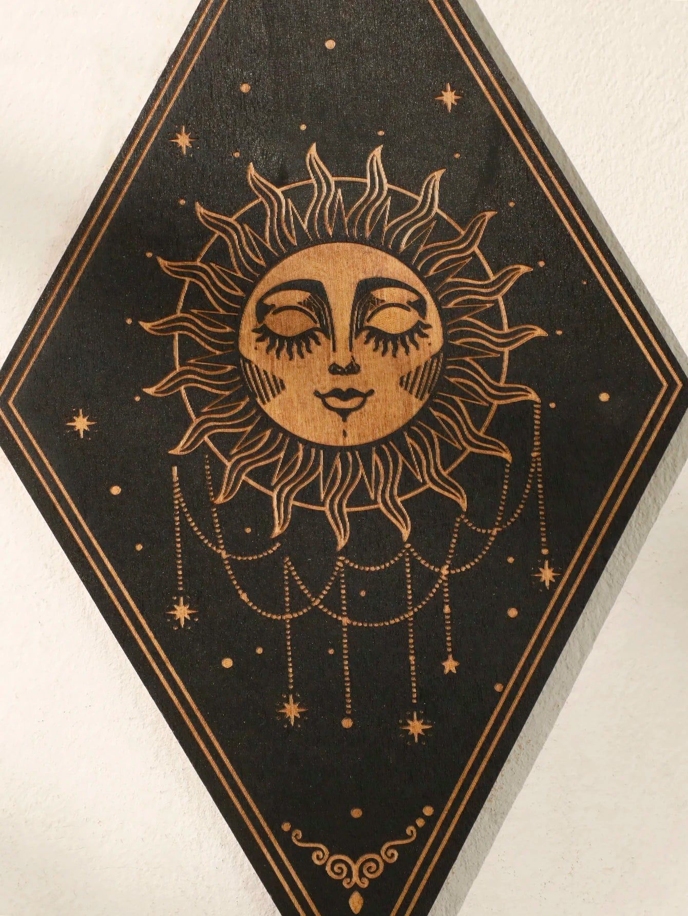A diamond-shaped black wall art piece featuring a gold-toned celestial design with a sun face, stars, and hanging dots, photographed against a light wall.