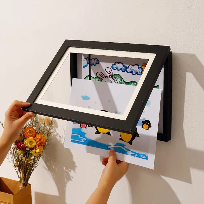 Person holding a black framed picture with children's drawings on a white wall.