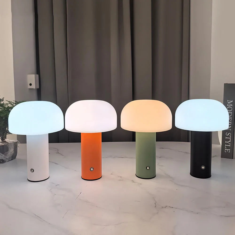 Four mushroom-shaped table lamps with white rounded shades and white, orange, green and black bases, illuminated on a pale tabletop.