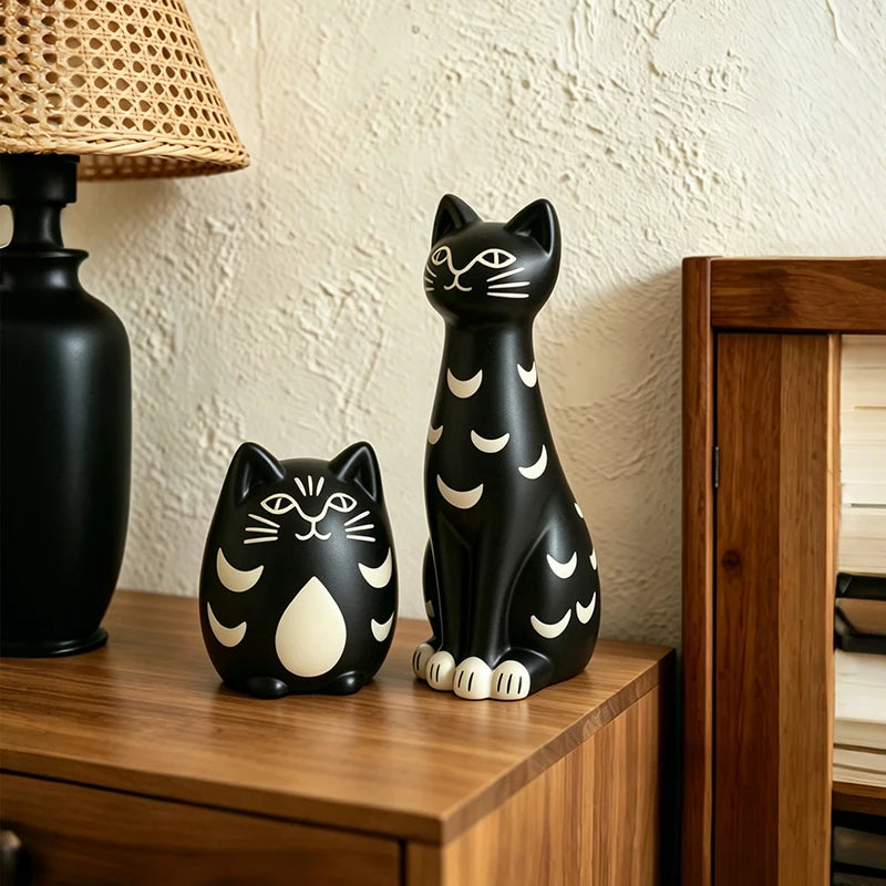 Two black cat figurines with white crescent and facial details displayed on a wooden side table next to a black lamp and textured wall.