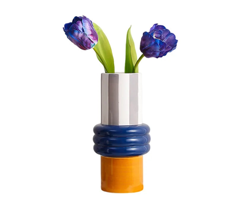 A modern sculptural ceramic vase with a white cylindrical top, a blue ribbed middle section, and an orange base, holding two purple tulips on a white background. The text 'IZZI Fish' is visible in the top-left corner.