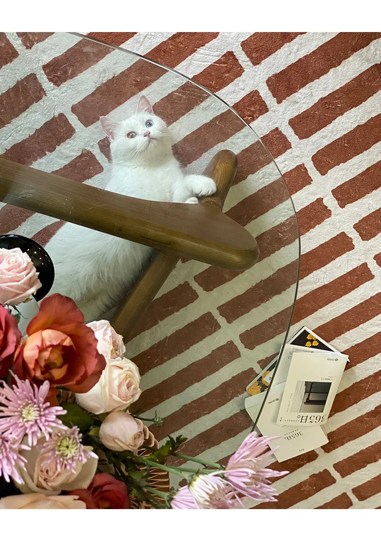 White cat on a glass table with flowers and a book underneath, against a brick wall.