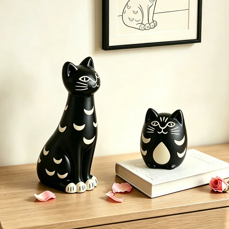 Two black cat-shaped decorative objects with white crescent moon patterns, one larger seated figure and one smaller bust, displayed on a wooden surface with a book, rose petals, and framed wall art in the background.