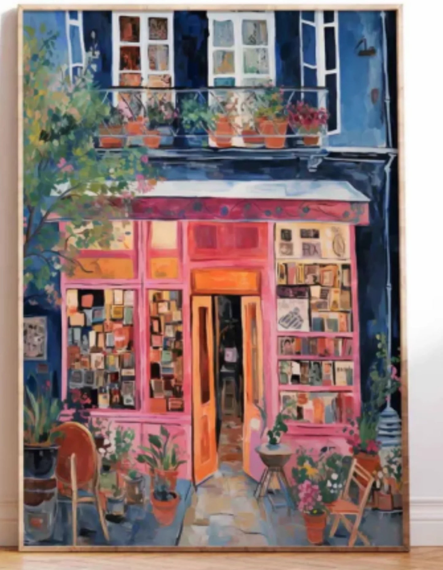 A framed painting depicting a colorful storefront with pink and orange shopfront, blue upper facade, windows, potted plants, chairs, and a light wood floor visible around the canvas.