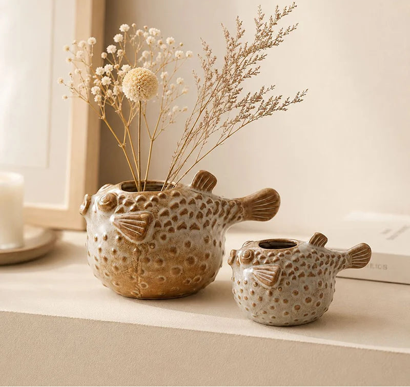 Two fish-shaped ceramic vases with a textured, spotted pattern in beige and brown, one filled with dried flowers, displayed on a shelf.